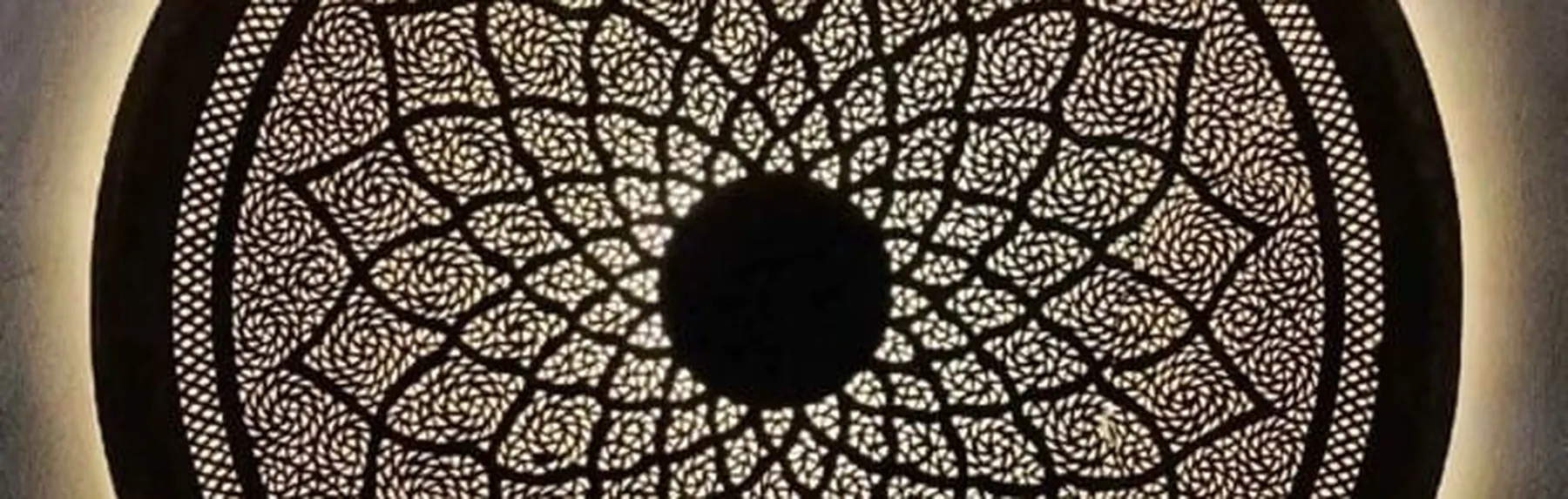 A round pierced brass wall light in a dense mandala pattern, backlit against a plaster wall