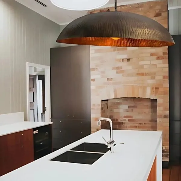 An oversized hammered brass dome pendant light hanging low above a kitchen island against a brick feature wall