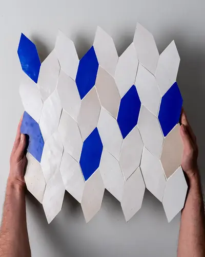 Hands holding a panel of white and blue diamond-shaped zellige tiles against a wall
