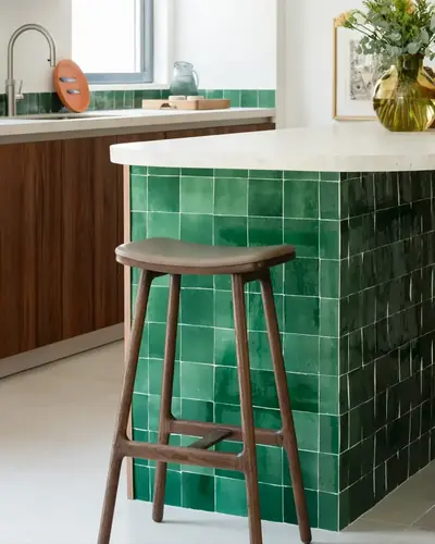 Emerald green zellige tiles cladding a kitchen island with a wooden stool alongside