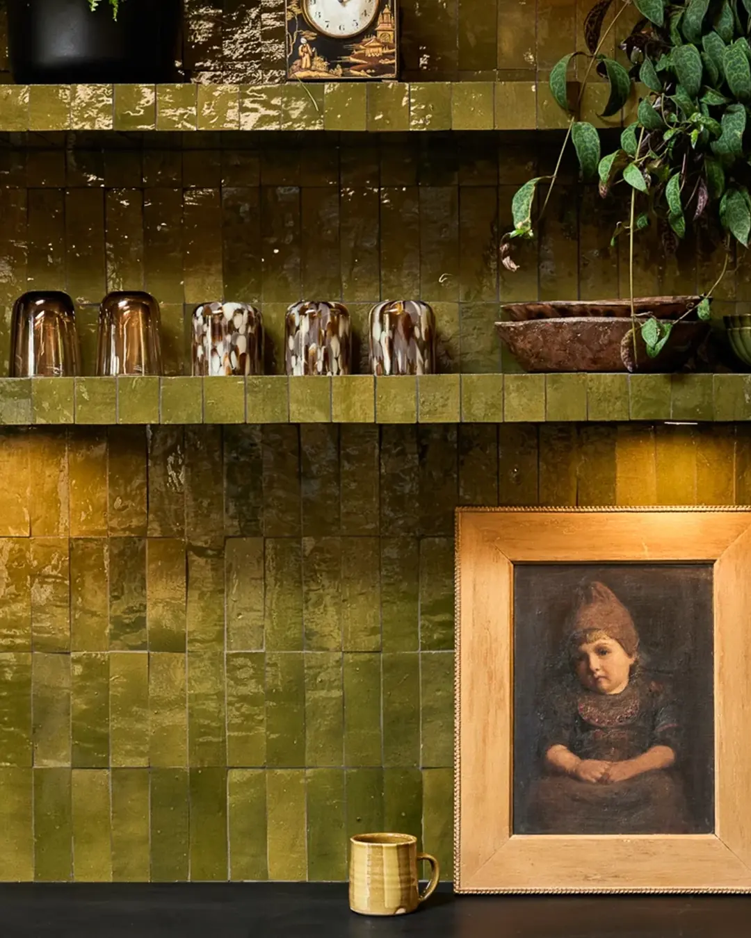 A deep olive green zellige wall behind open kitchen shelving, the joints reading as a close tone rather than a contrasting grid