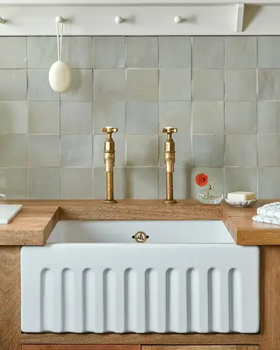 Pale green zellige wall above a white fireclay butler sink with brass taps