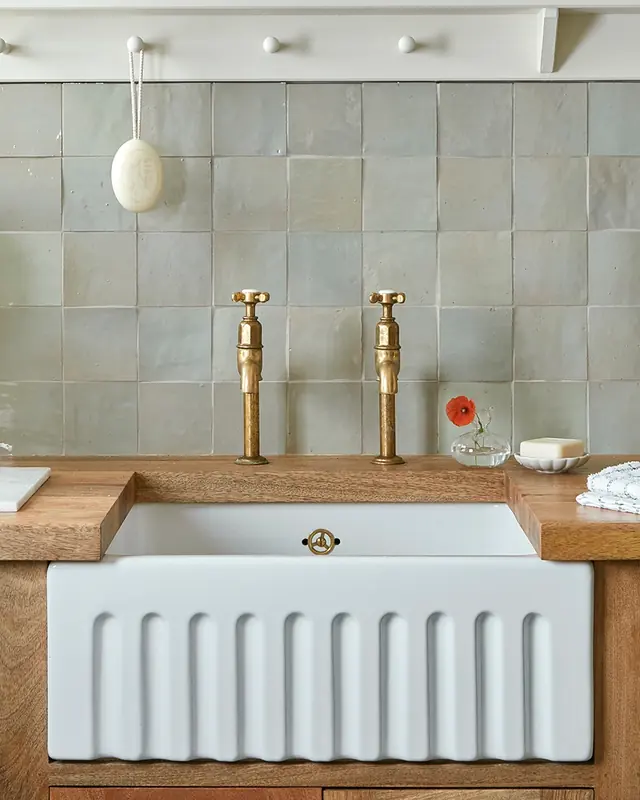 A pale green zellige wall above a white fireclay sink with brass taps