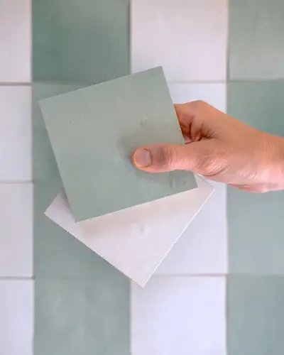 A hand holding a pale green zellige sample tile against a tiled wall for comparison
