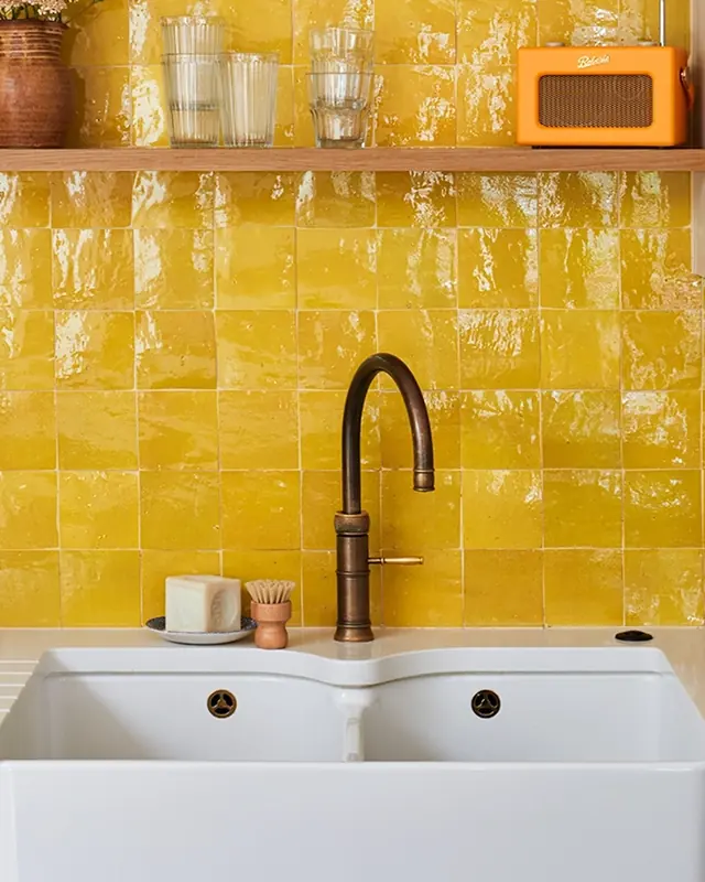 Saffron yellow zellige splashback behind a white sink and brass tap